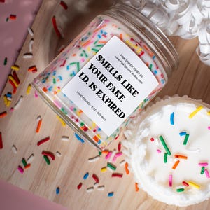 May include: A white candle with colorful sprinkles on top in a glass jar. The label reads "Smells Like Your Fake I.D. Is Expired."  The candle is on a wooden surface with more sprinkles scattered around it.