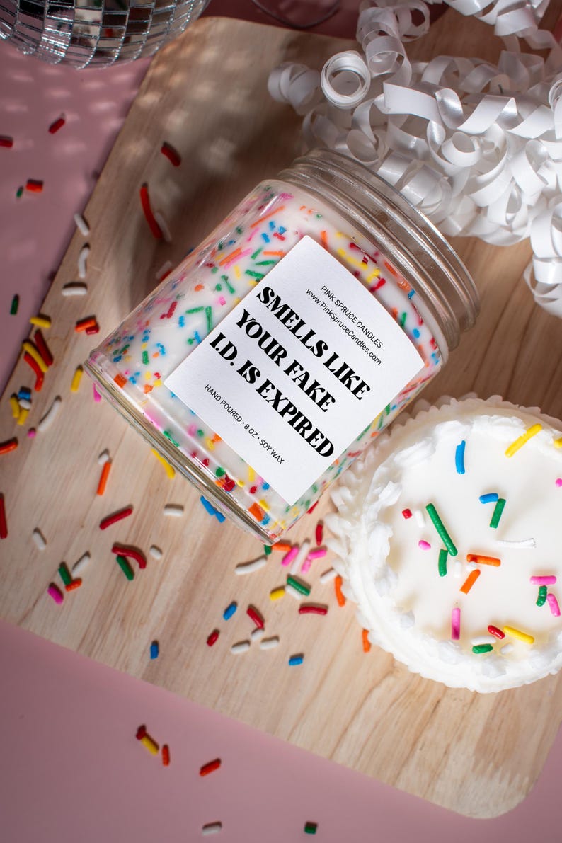 May include: A white candle with colorful sprinkles on top in a glass jar. The label reads "Smells Like Your Fake I.D. Is Expired."  The candle is on a wooden surface with more sprinkles scattered around it.