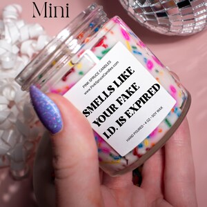 May include: A small glass jar candle with a white label that says "Smells Like Your Fake I.D. Is Expired". The candle is decorated with colorful sprinkles and the label also includes the website address "www.PinkSpruceCandles.com".