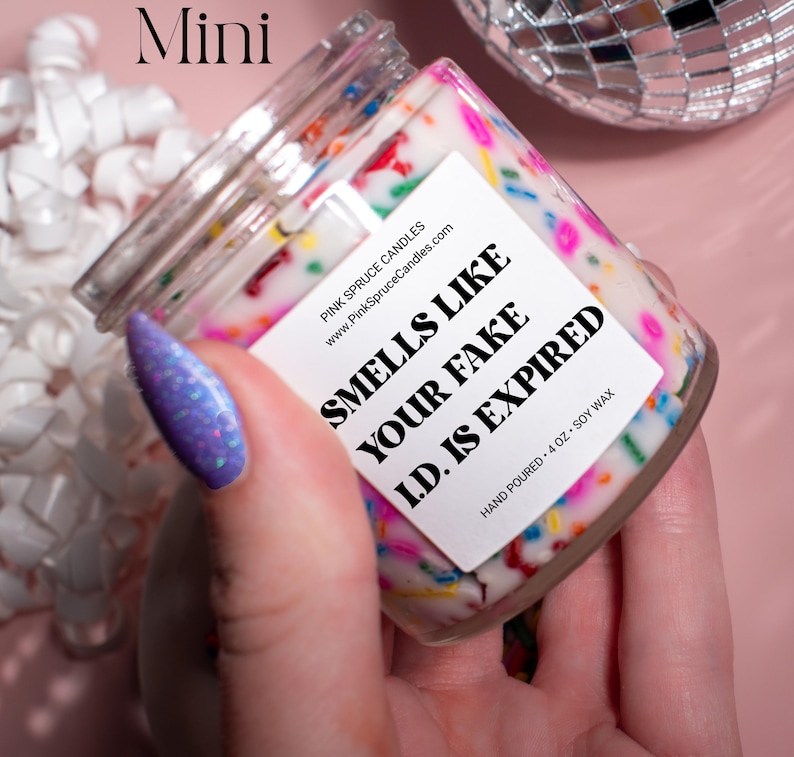 May include: A small glass jar candle with a white label that says "Smells Like Your Fake I.D. Is Expired". The candle is decorated with colorful sprinkles and the label also includes the website address "www.PinkSpruceCandles.com".