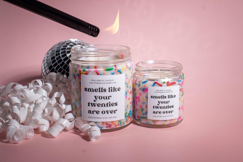 May include: Two candles with a white background and colorful sprinkles. The candles have labels that say "smells like your twenties are over".