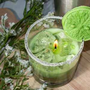 May include: A green candle in a glass jar with a sugared rim. The candle is topped with a green lime slice and ice cubes.