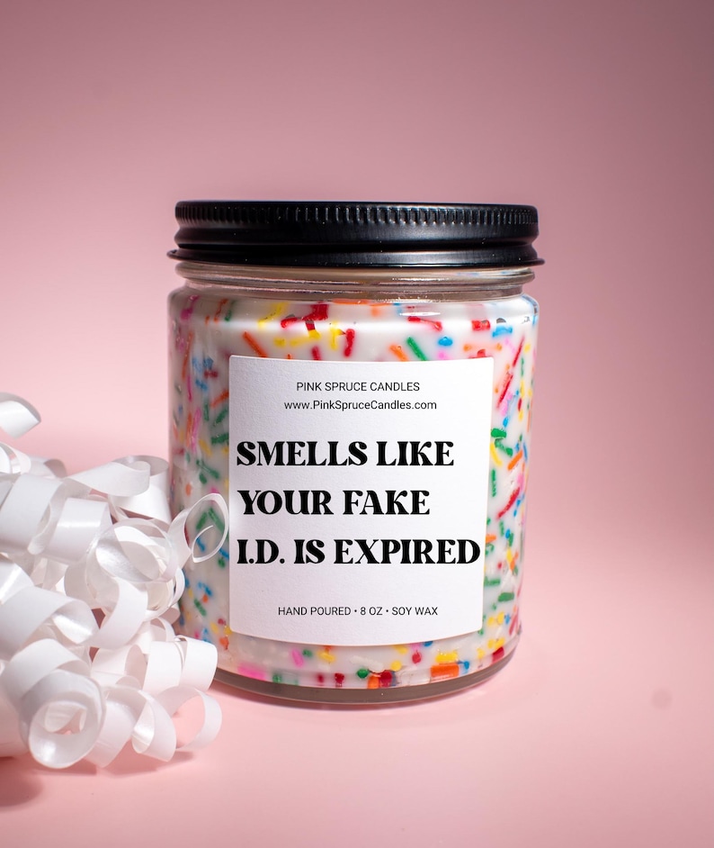 May include: A white candle jar with a black lid and a colorful sprinkle design. The label reads "Smells Like Your Fake I.D. Is Expired" and "Pink Spruce Candles". The candle is hand-poured and made with soy wax.