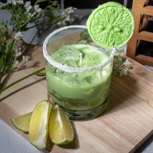 May include: A green candle in a glass jar with a sugared rim. The candle is shaped like a lime and is topped with a lime slice. There are lime wedges on a wooden cutting board.