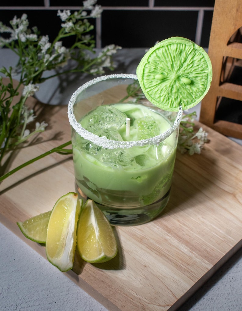 May include: A green candle in a glass jar with a sugared rim. The candle is shaped like a lime and is topped with a lime slice. There are lime wedges on a wooden cutting board.