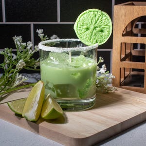 May include: A green cocktail with ice in a glass rimmed with white sugar. A green lime wedge is on a stick in the drink. There are lime wedges on a wooden cutting board in front of the glass.