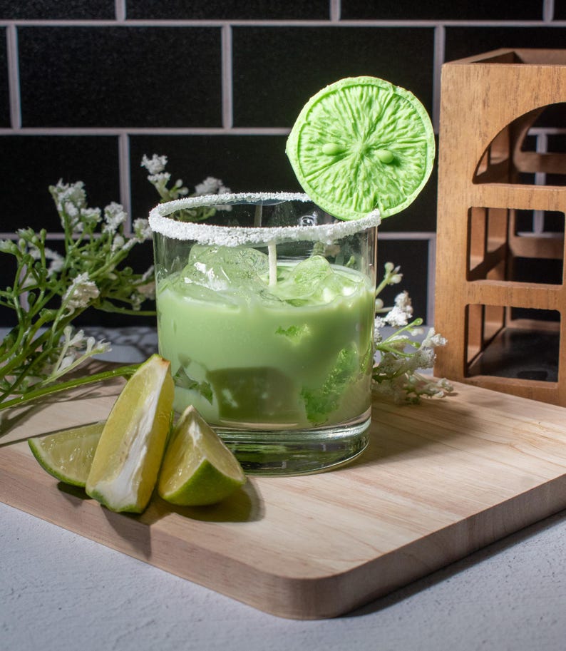 May include: A green cocktail with ice in a glass rimmed with white sugar. A green lime wedge is on a stick in the drink. There are lime wedges on a wooden cutting board in front of the glass.