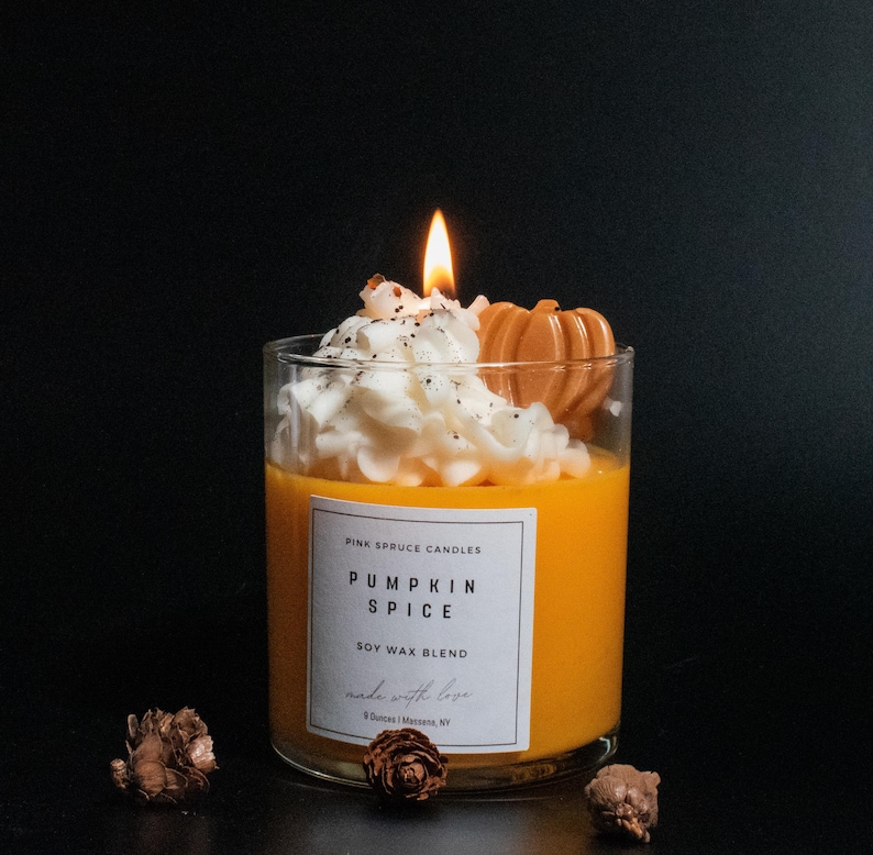 May include: A glass jar candle with a white whipped cream topping and a small orange pumpkin decoration. The candle is labeled "Pumpkin Spice Soy Wax Blend" and "Pink Spruce Candles".