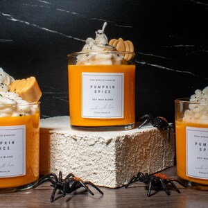 May include: Three orange pumpkin spice soy wax blend candles with whipped cream and caramel topping. The candles are on a white and brown surface with black spiders.