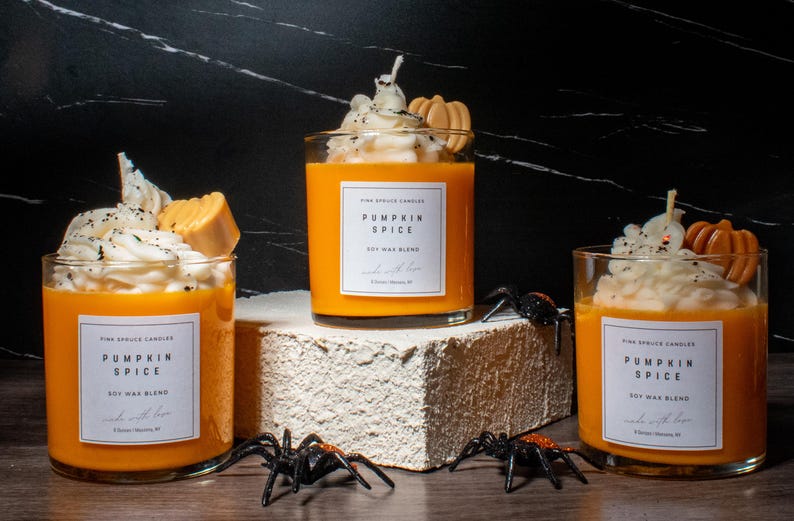 May include: Three orange pumpkin spice soy wax blend candles with whipped cream and caramel topping. The candles are on a white and brown surface with black spiders.
