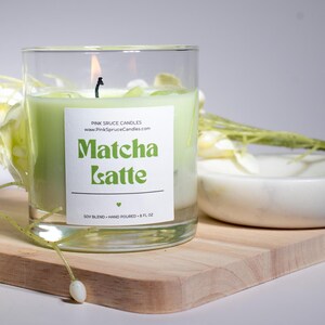 May include: A green soy candle in a clear glass jar with a white label that reads "Matcha Latte" and "Pink Spruce Candles". The candle is on a wooden surface with a white marble dish and white flowers.