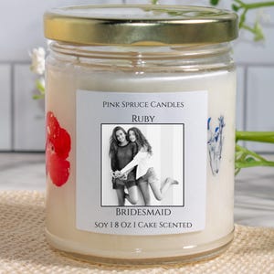 Może przedstawiać: Świeca z białego wosku sojowego w przezroczystym szklanym słoiku ze złotą pokrywką. Etykieta zawiera napis Pink Spruce Candles, Ruby, Bridesmaid, Soy 8 oz Cake Scented oraz czarno-białe zdjęcie dw&oacute;ch os&oacute;b. Widoczne są czerwone i niebieskie kwiaty.