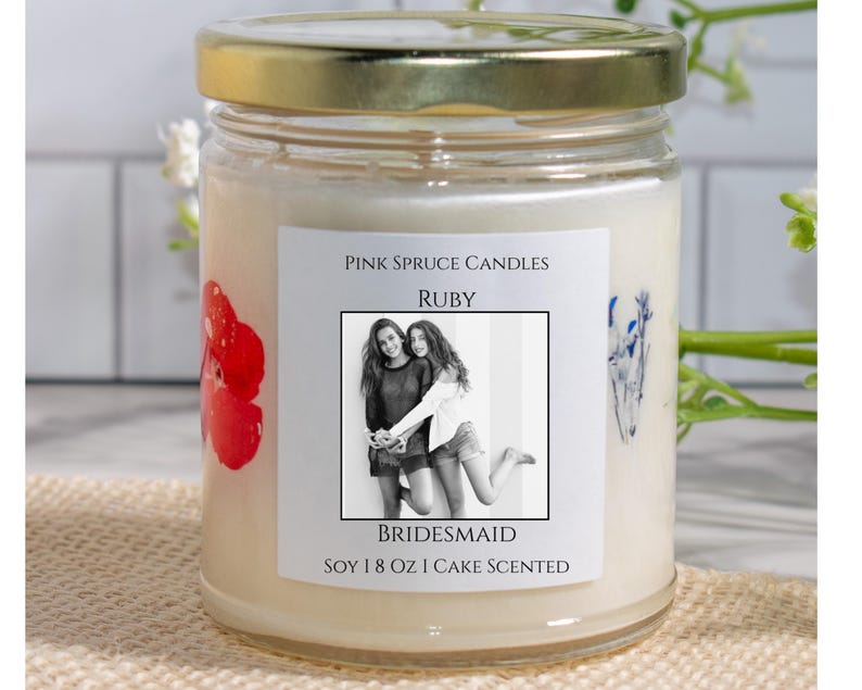 Może przedstawiać: Świeca z białego wosku sojowego w przezroczystym szklanym słoiku ze złotą pokrywką. Etykieta zawiera napis Pink Spruce Candles, Ruby, Bridesmaid, Soy 8 oz Cake Scented oraz czarno-białe zdjęcie dw&oacute;ch os&oacute;b. Widoczne są czerwone i niebieskie kwiaty.