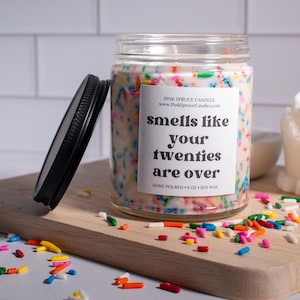 May include: A white candle with colorful sprinkles on top in a clear glass jar with a black lid. The label on the jar reads "smells like your twenties are over" and "Pink Spruce Candles".