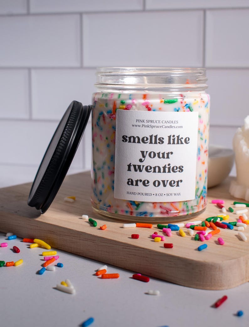 May include: A white candle with colorful sprinkles on top in a clear glass jar with a black lid. The label on the jar reads "smells like your twenties are over" and "Pink Spruce Candles".