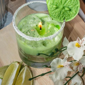 May include: A green candle in a glass jar with a lime wedge on top. The candle is surrounded by ice cubes and a white rim of sugar. The jar is sitting on a wooden surface with white flowers.