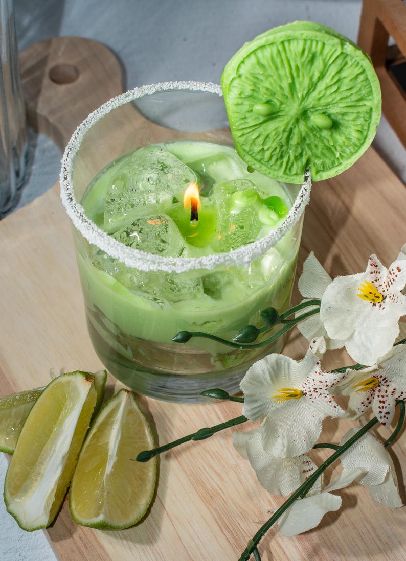May include: A green candle in a glass jar with a lime wedge on top. The candle is surrounded by ice cubes and a white rim of sugar. The jar is sitting on a wooden surface with white flowers.