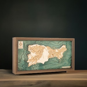 Könnte beinhalten: Eine Holzkarte der Insel Capri, Italien, mit einer Kompassrose und dem Text "Isola di Capri" in der linken unteren Ecke. Die Karte ist in einem dunklen Holzrahmen gerahmt und hat einen blaugrünen Hintergrund.