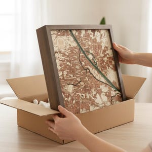 Op de afbeelding: Een vierkant wandkunstwerk met een donkerhouten lijst, voorzien van een gedetailleerde, meerlaagse topografische kaart in tinten bruin, crème en groen. De kaart toont complexe stratenpatronen en een opvallende groene rivierloop.