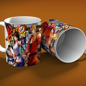 May include: White ceramic mug featuring the text "ART OF FIGHTING" and illustrations of video game characters. The mug has a white handle and a colorful design, with a second mug visible in the background.