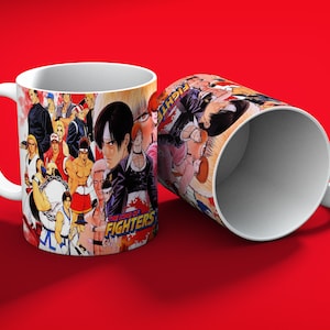 May include: Two white ceramic mugs with a colorful graphic design featuring characters from "The King of Fighters" video game. The mugs are set against a red background, with one mug upright and the other tilted on its side.