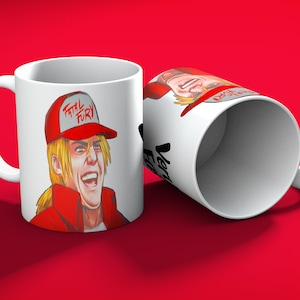 May include: White ceramic mug featuring a cartoon illustration of a character from the video game "Fatal Fury." The character is depicted with blonde hair, a red hat with the text "Fatal Fury," and a red jacket. The mug is on a red background.