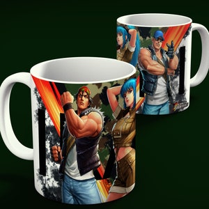 May include: White ceramic mug featuring illustrations of characters from "The King of Fighters" video game. The design includes a camouflage pattern, bold graphics, and the game's logo. The mug has a white handle and interior.