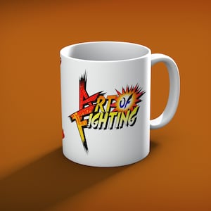 May include: White ceramic mug with the text "ART OF FIGHTING" in a colorful, comic book style design. The mug has a curved handle and is set against an orange background. The text is in red, yellow, and blue.