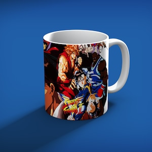 May include: White ceramic mug featuring a colorful graphic of characters from the video game Street Fighter II. The mug has a white handle and a blue background.