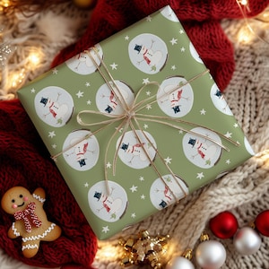 May include: A wrapped gift with a festive snowman pattern on green paper, tied with gold ribbon. The wrapping paper features repeating circular designs of snowmen wearing top hats and scarves, interspersed with white stars. A gingerbread man and Christmas ornaments are in the background.