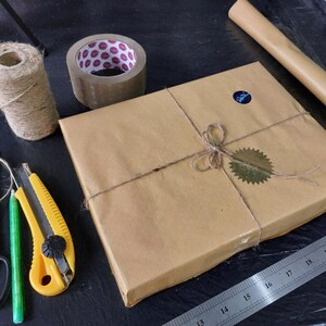 May include: A brown package wrapped in twine and sealed with a gold sticker. The package is sitting on a black surface with a ruler, scissors, a utility knife, and a roll of brown packing tape.