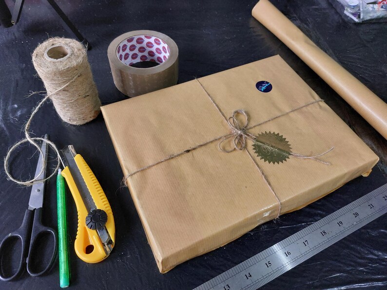 May include: A brown package wrapped in twine and sealed with a gold sticker. The package is sitting on a black surface with a ruler, scissors, a utility knife, and a roll of brown packing tape.
