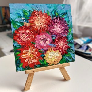 May include: A small painting on a wooden easel depicts a bouquet of colorful flowers. The flowers are painted in a vibrant palette of red, pink, yellow, and purple. The background is a light blue.