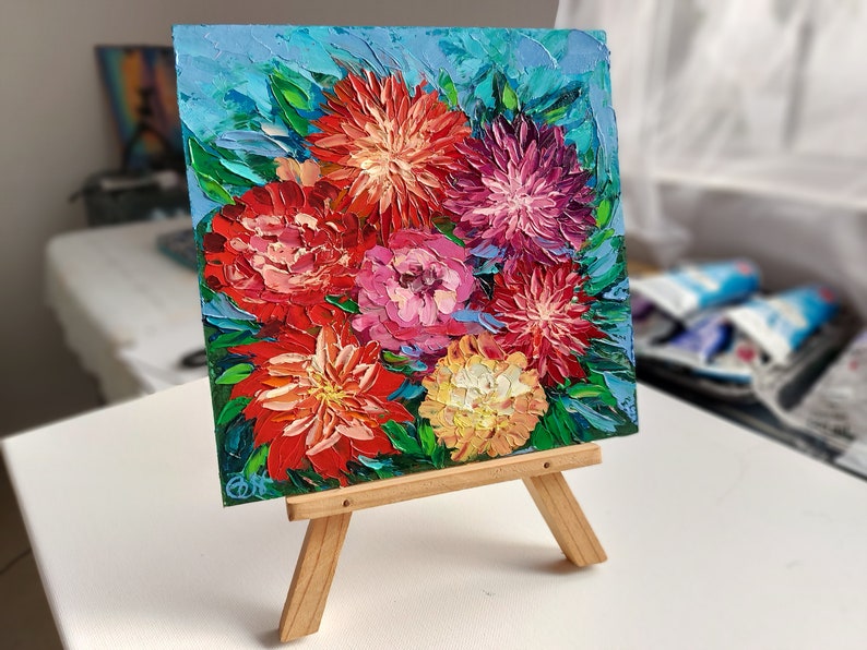 May include: A small painting on a wooden easel depicts a bouquet of colorful flowers. The flowers are painted in a vibrant palette of red, pink, yellow, and purple. The background is a light blue.