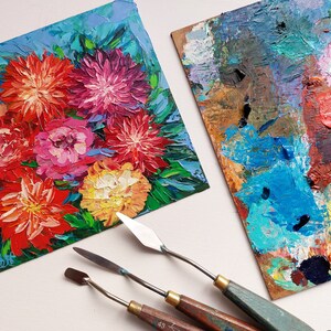 May include: A close-up of a small painting of a bouquet of colorful flowers, with a blue background. Three painting knives are also in the image, along with a palette with various colors of paint.