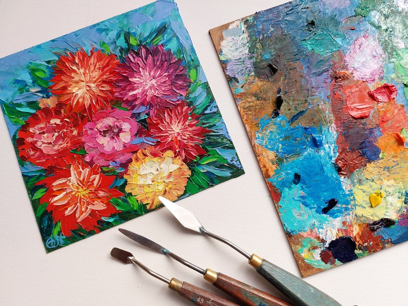 May include: A close-up of a small painting of a bouquet of colorful flowers, with a blue background. Three painting knives are also in the image, along with a palette with various colors of paint.