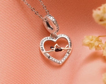 Puede incluir: Un collar plateado con un colgante en forma de corazón. El colgante presenta un diseño de nadador y el texto grabado "She believed she could so she did". El corazón está bordeado con cristales transparentes.