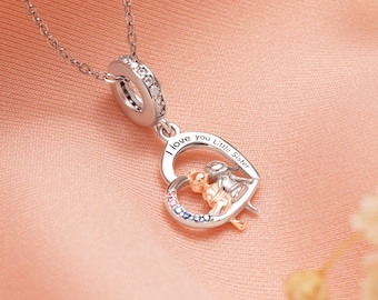 Puede incluir: Un collar plateado con un colgante en forma de corazón. El colgante tiene las palabras "I love you Little Sister" y presenta una pequeña figura. El collar está sobre una superficie rosa.