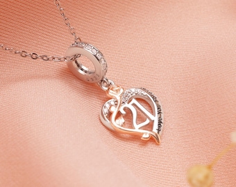 Puede incluir: Collar de plata con un colgante en forma de corazón. El colgante presenta un contorno de oro rosa, el número 21 y pequeñas piedras transparentes. El collar está sobre una superficie de satén rosa.