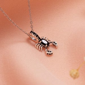 May include: A silver-toned necklace featuring a scorpion pendant. The pendant has a black oval-shaped center stone and black accent stones. The scorpion's body is detailed with small claws and a curved tail. The necklace chain is delicate and silver.