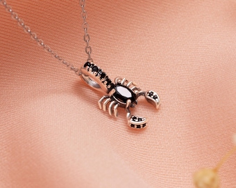 Puede incluir: Un collar plateado con un colgante de escorpión. El colgante tiene una piedra central negra de forma ovalada y piedras negras de acento. El cuerpo del escorpión está detallado con pequeñas pinzas y una cola curvada.