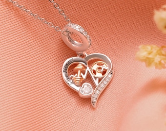 Puede incluir: Un collar plateado con un colgante en forma de corazón. El colgante presenta una mancuerna, una placa de pesas y la palabra "Fitness". El corazón está adornado con pequeñas piedras transparentes y una pequeña piedra en forma de corazón.