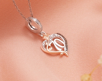 Puede incluir: Un collar plateado con un colgante en forma de corazón. El colgante presenta un contorno de oro rosa, incrustado con pequeñas piedras transparentes, y las palabras "Love You Forever". El collar se muestra sobre una tela rosa.