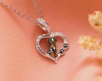Puede incluir: Collar plateado con un colgante en forma de corazón. El colgante presenta una pequeña figura de perro, huellas de patas y las palabras "I love you forever". El collar se muestra sobre una superficie rosa.