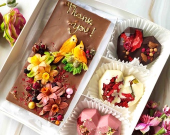 Puede incluir: Una caja de regalo blanca con una barra de chocolate decorada con flores y pájaros. La caja también contiene cuatro corazones de chocolate, dos de chocolate blanco y dos de chocolate negro. Los corazones de chocolate están decorados con frutos secos y nueces. La barra de chocolate tiene las palabras "Thank you!" escritas en ella.