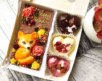 Puede incluir: Una caja de regalo blanca que contiene una barra de chocolate con un diseño de zorro y flores. La caja también incluye tres chocolates con forma de corazón, uno con relleno de chocolate y malvavisco, otro con relleno de chocolate blanco y almendras, y otro con relleno de chocolate rosa y almendras. La barra de chocolate tiene las palabras "Happy Birthday" escritas en ella.