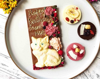 Puede incluir: Una barra de chocolate decorada con un gatito de chocolate blanco, flores y chispas. La barra tiene las palabras "Happy Birthday from Pixie" escritas en dorado.