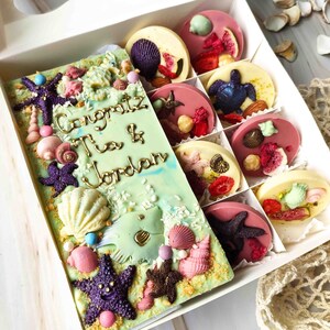 May include: A white box filled with assorted chocolate discs decorated with various toppings, including fruit, nuts, and seashells. A larger chocolate bar with a green background and gold lettering reads "Congrats Tia & Jordan".
