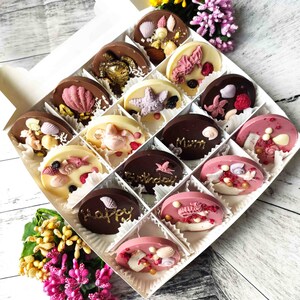 May include: A white box filled with assorted chocolate treats. The chocolates are decorated with various designs, including seashells, starfish, and the word "Happy".