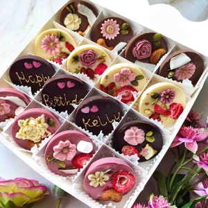 May include: A white box filled with assorted chocolate treats. The chocolates are decorated with various designs, including flowers, hearts, and the words "Happy Birthday" and "Kelly".